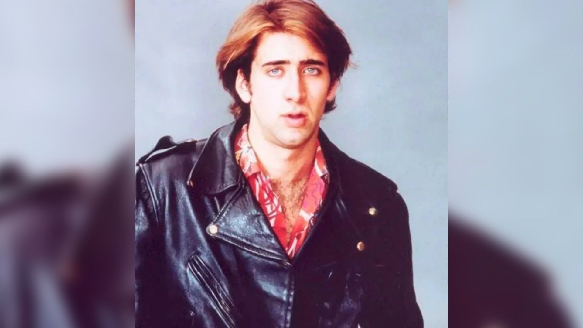 A young man with thick hair in a leather jacket and a bright shirt looks at the camera against a gray wall
