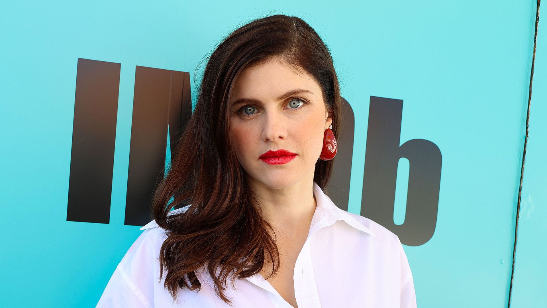 Actriz Alexandra Daddario en una blusa blanca contra un fondo turquesa