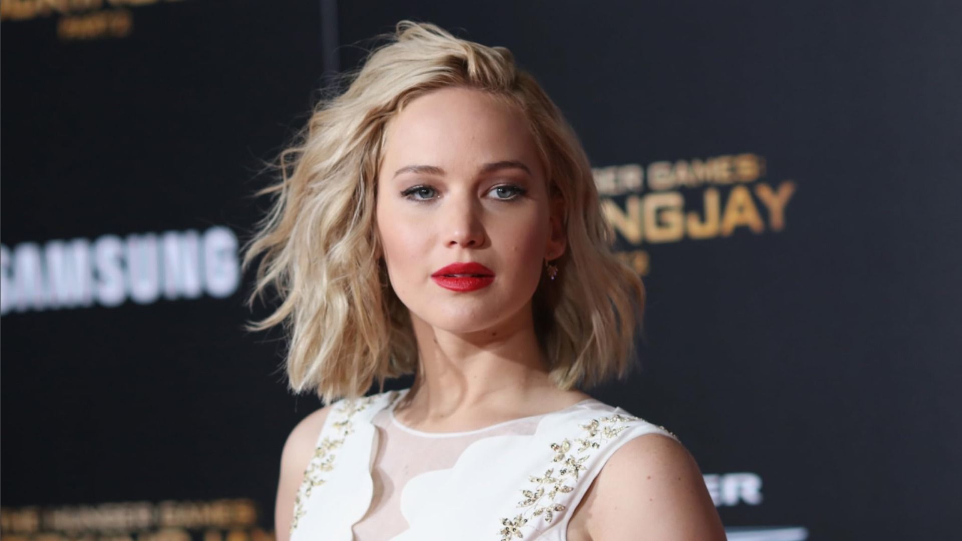 Actriz Jennifer Lawrence mirando hacia un lado con un vestido blanco contra el fondo del cartel de la película 'The Hunger Games'