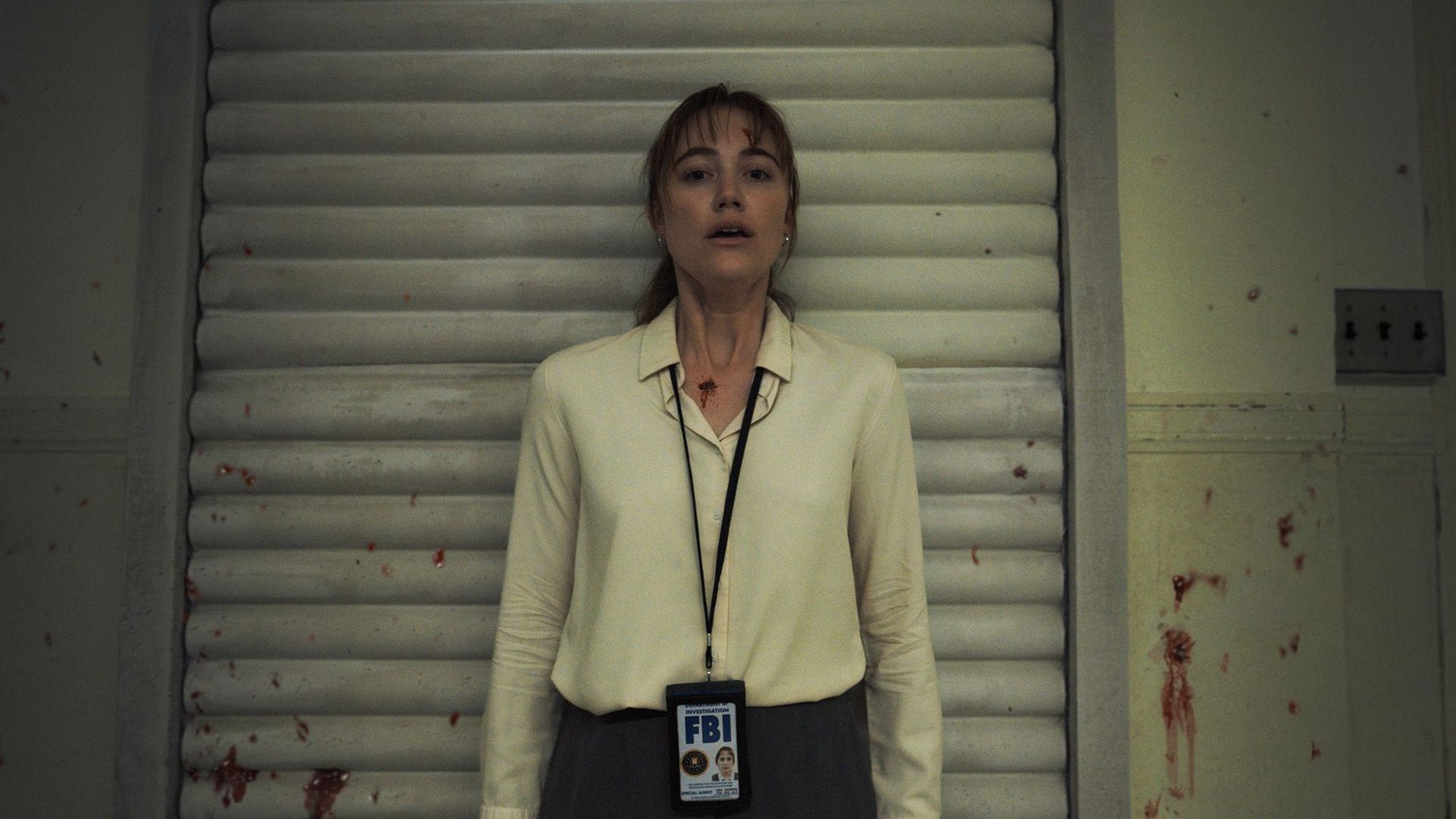 Maika Monroe con una camisa blanca y una placa del FBI alrededor de su cuello, con una expresión aterrorizada contra la pared, rodeada de salpicaduras de sangre