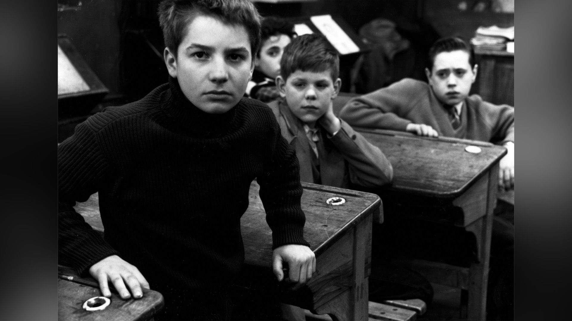 Un aula escolar, varios chicos sentados en sus pupitres, uno de ellos mirando seriamente a la cámara