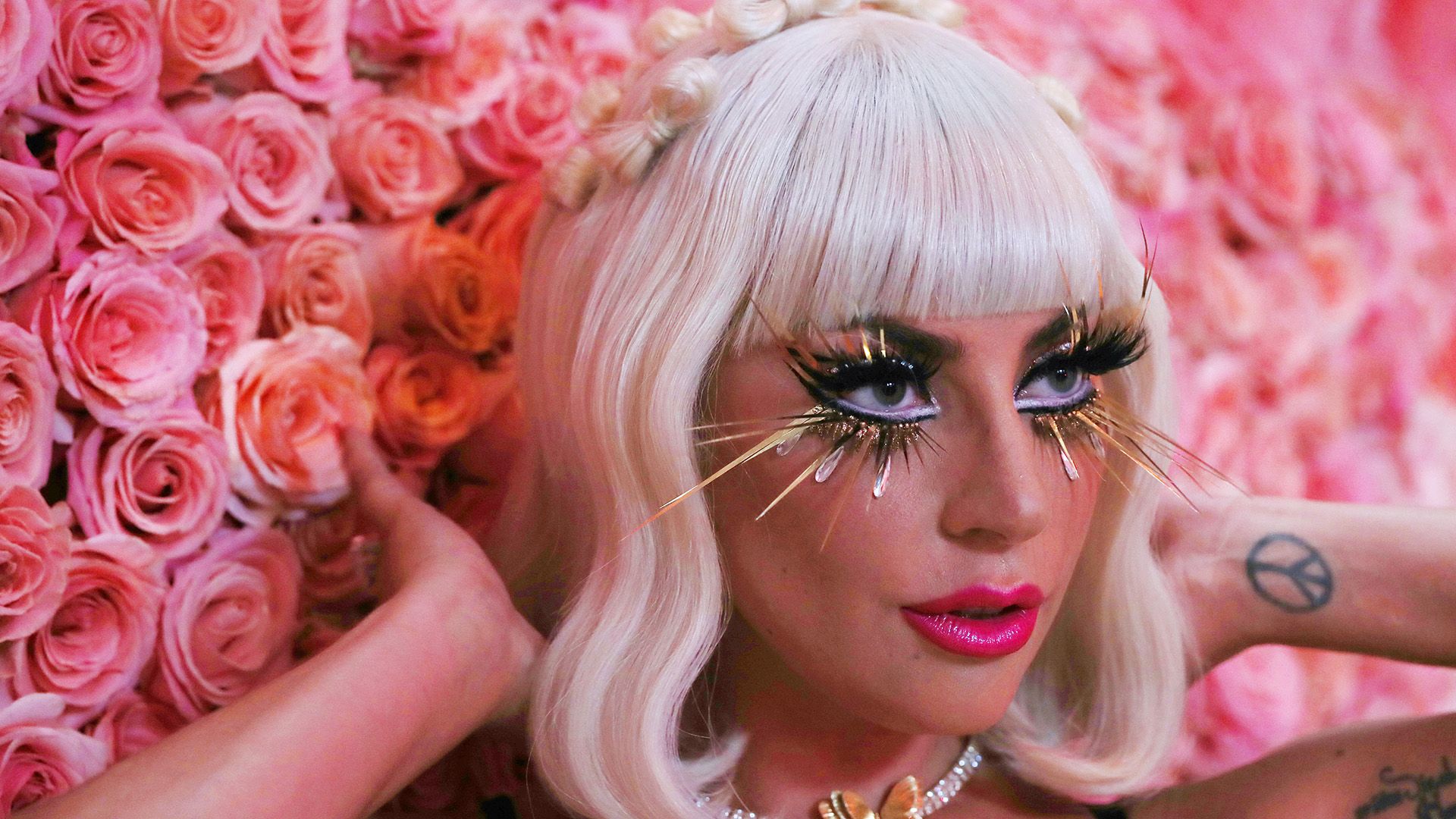 Lady Gaga con maquillaje llamativo y una peluca blanca contra un fondo de flores rosadas