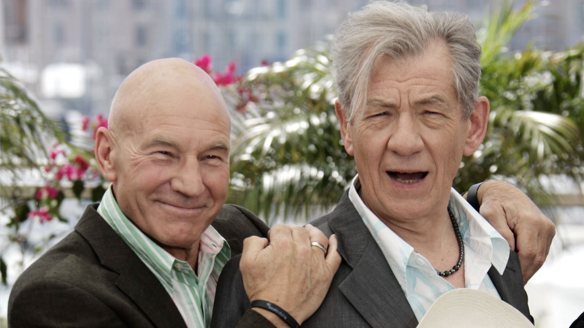 El actor Patrick Stewart sonríe y abraza al actor Ian McKellen sobre un fondo de flores