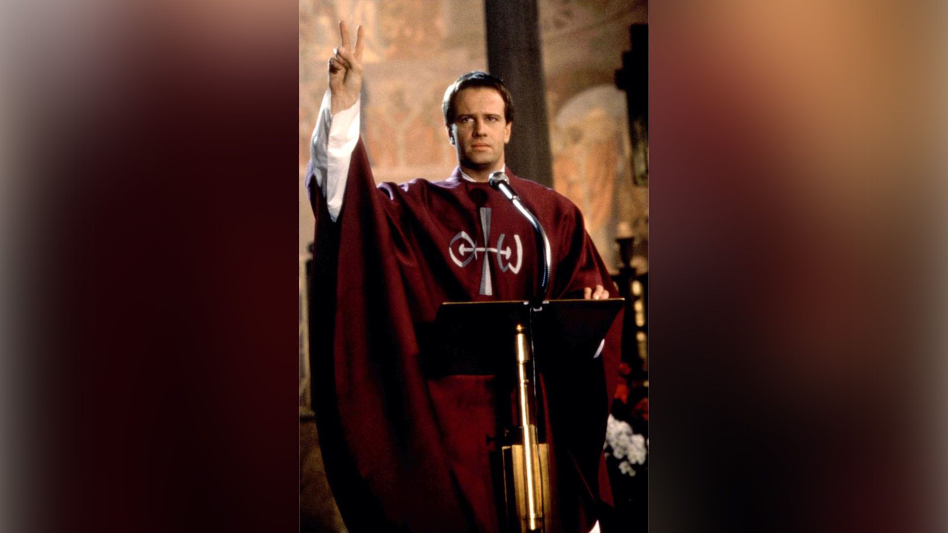 Christopher Lambert con una túnica roja de sacerdote se encuentra detrás del podio en la iglesia