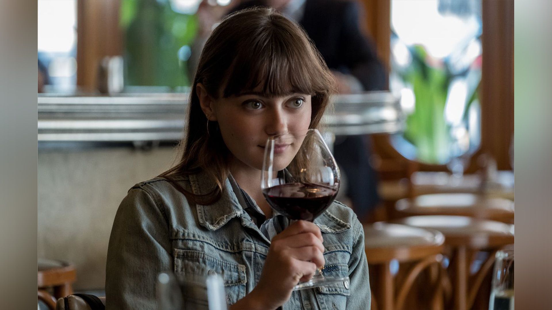 Ella Purnell con una chaqueta de mezclilla sosteniendo un vaso con un líquido rojo oscuro frente a su cara