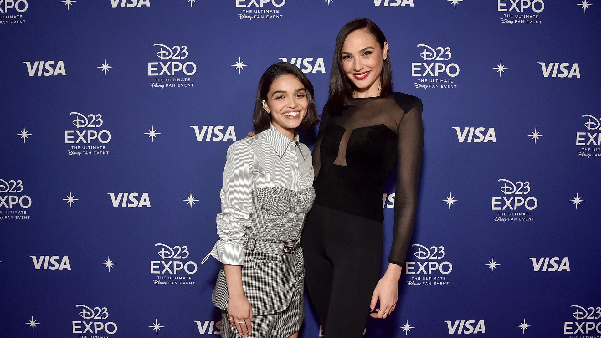 Rachel Zegler en ropa clara y Gal Gadot en negro contra un fondo azul y ríen