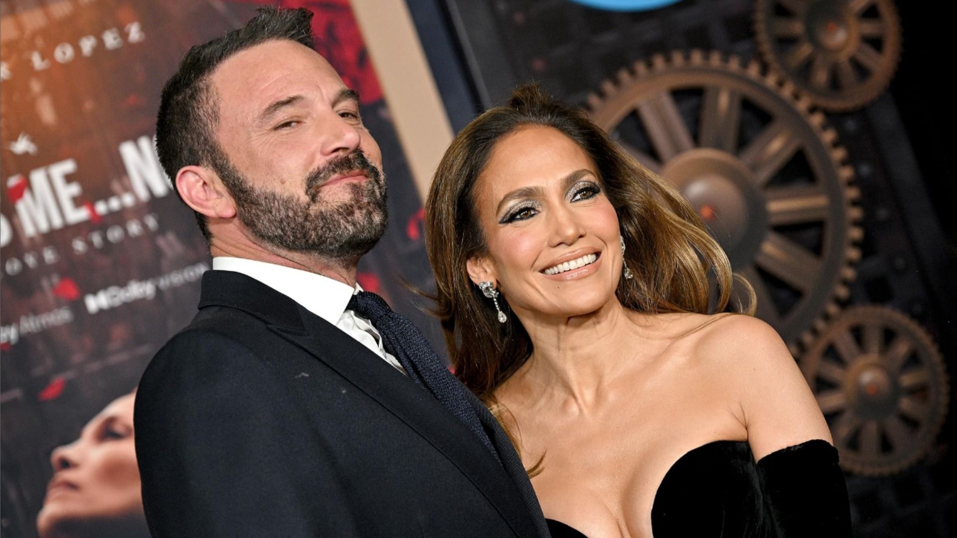 El actor Ben Affleck con un traje negro y la actriz Jennifer Lopez con un vestido negro sonríen frente a un póster borroso de la película 'It's Me. Now'