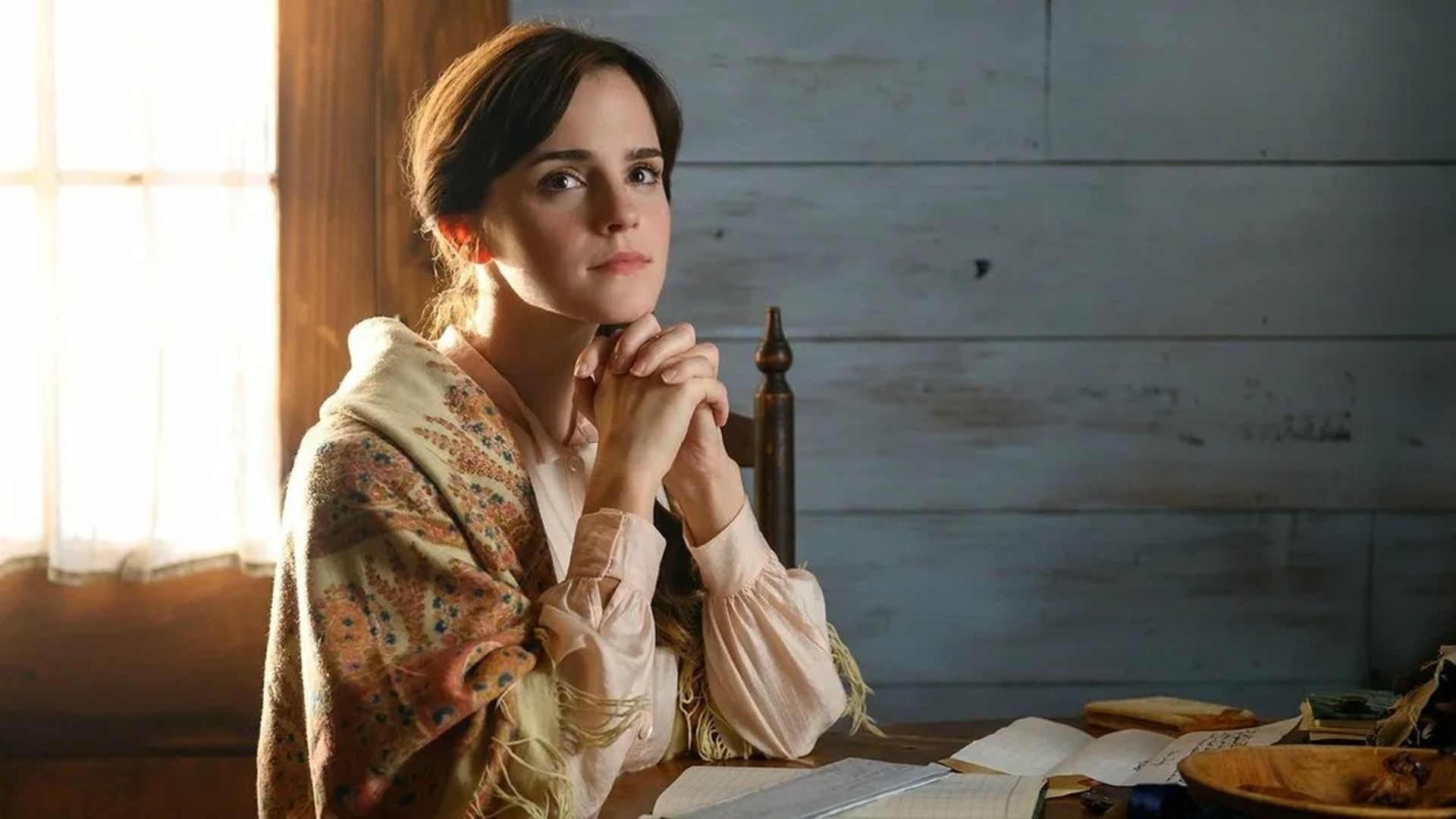 Emma Watson sentada a la mesa con una expresión pensativa en su rostro, con las manos juntas
