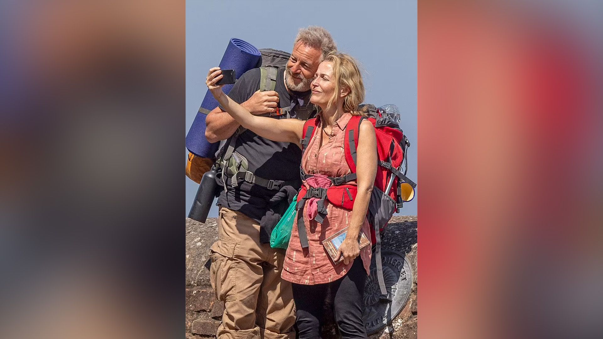 Jason Isaacs y Gillian Anderson con mochilas en sus espaldas están tomando una selfie en el teléfono
