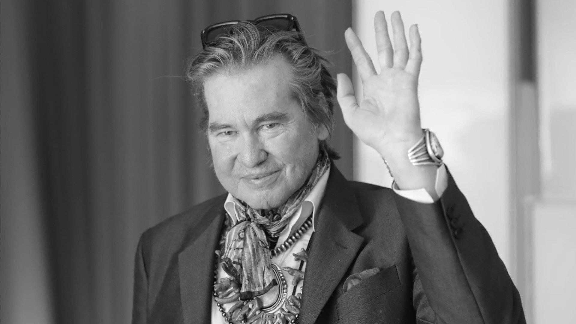 Actor Val Kilmer in a costume and with glasses on his head smiles and waves at the camera
