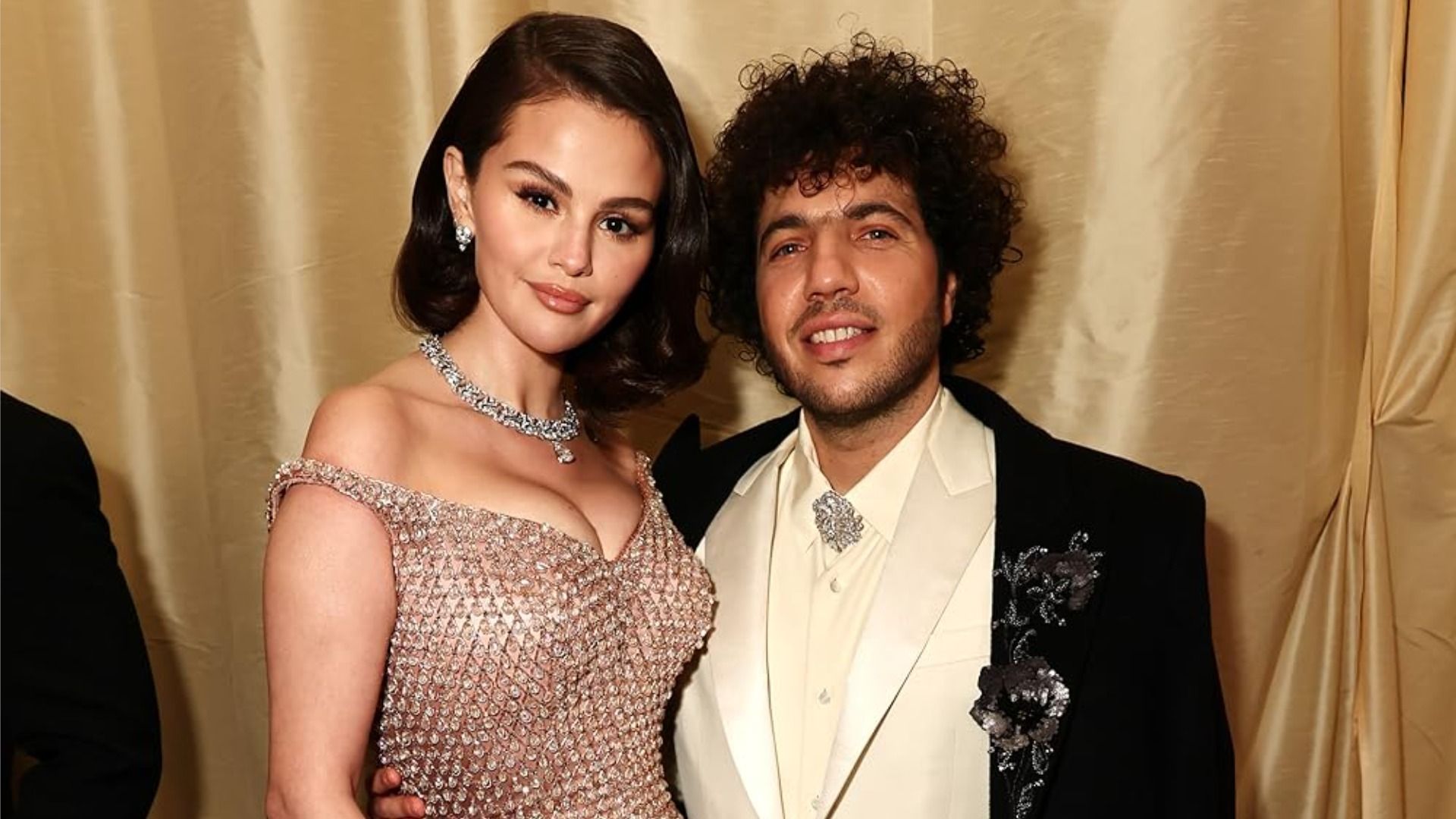 La actriz Selena Gomez y su prometido, el productor musical Benny Blanco, están abrazados y sonriendo a la cámara contra un fondo dorado