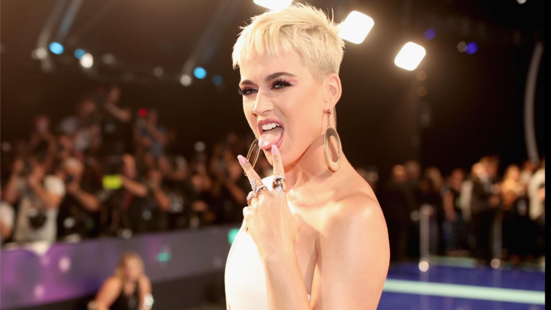 La cantante Katy Perry saca la lengua y levanta dos dedos con un vestido blanco contra el fondo del público