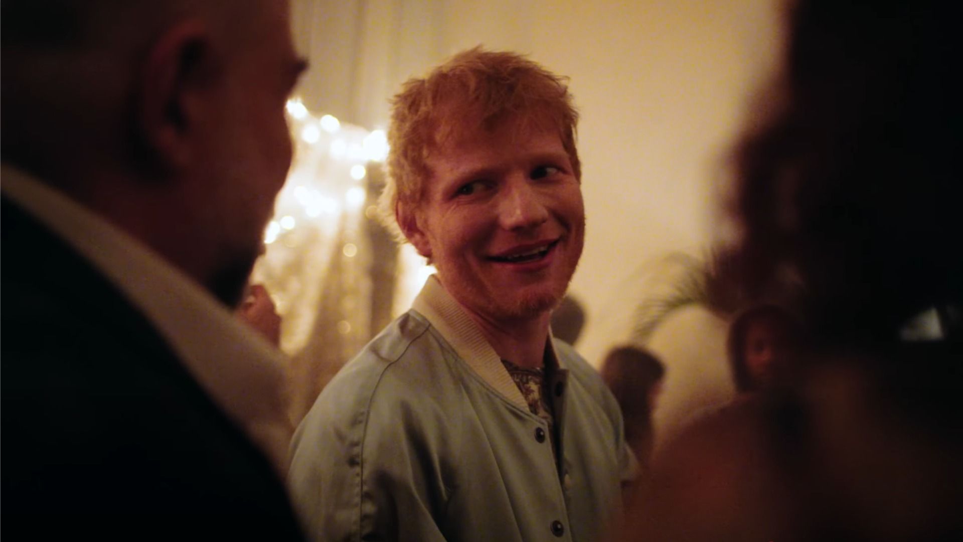 El cantante Ed Sheeran mira y sonríe en la luz amarillo-naranja junto a las figuras de personas