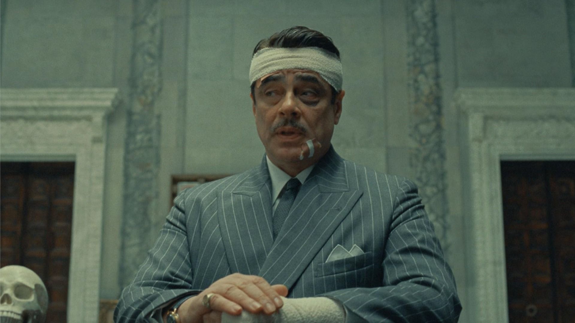 Actor Benicio del Toro en una chaqueta gris y con la cabeza vendada sentado en una mesa contra una pared