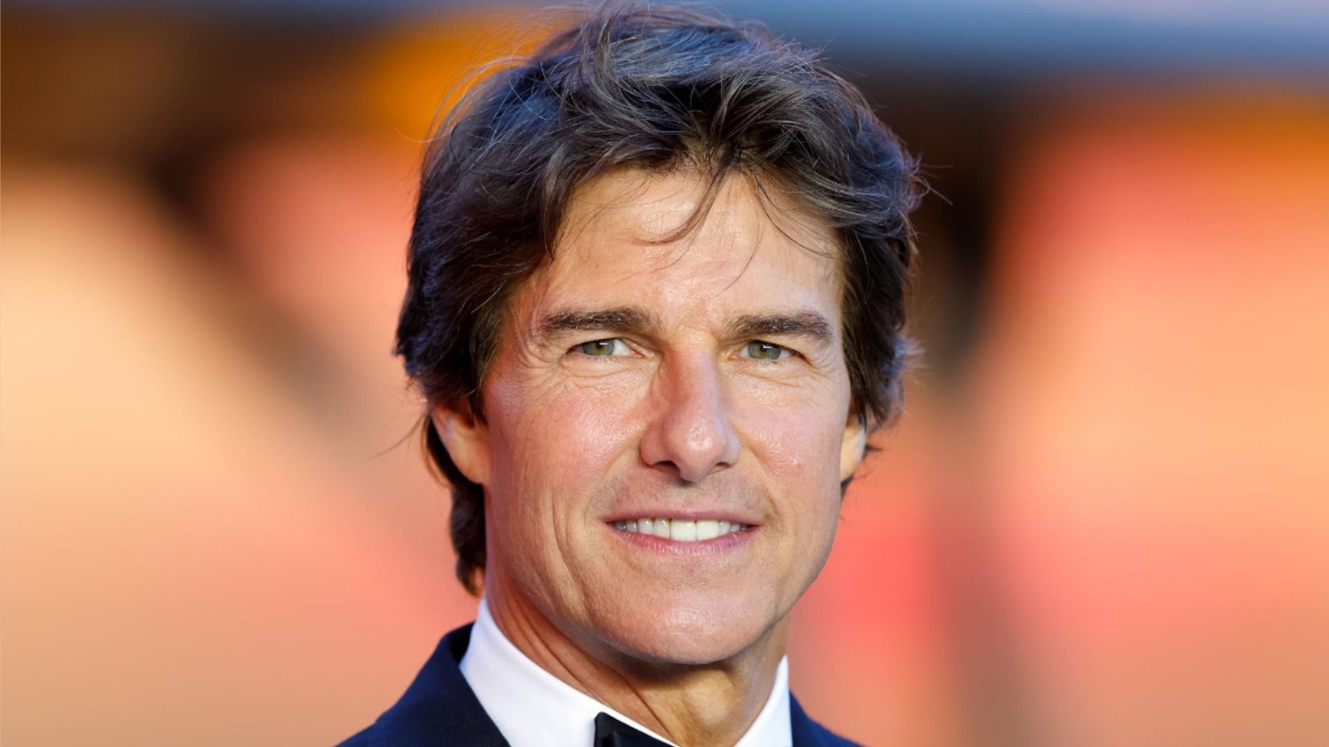 El actor Tom Cruise posa con un traje clásico y sonríe a la cámara contra un fondo desenfocado