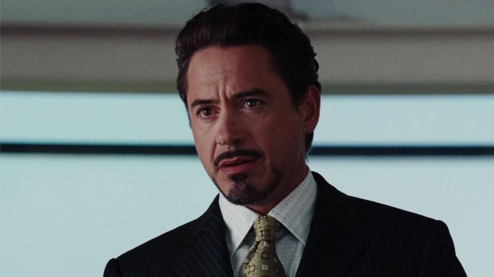 Actor Robert Downey Jr. con bigote y barba en un traje clásico mirando hacia un lado contra el fondo de una ventana