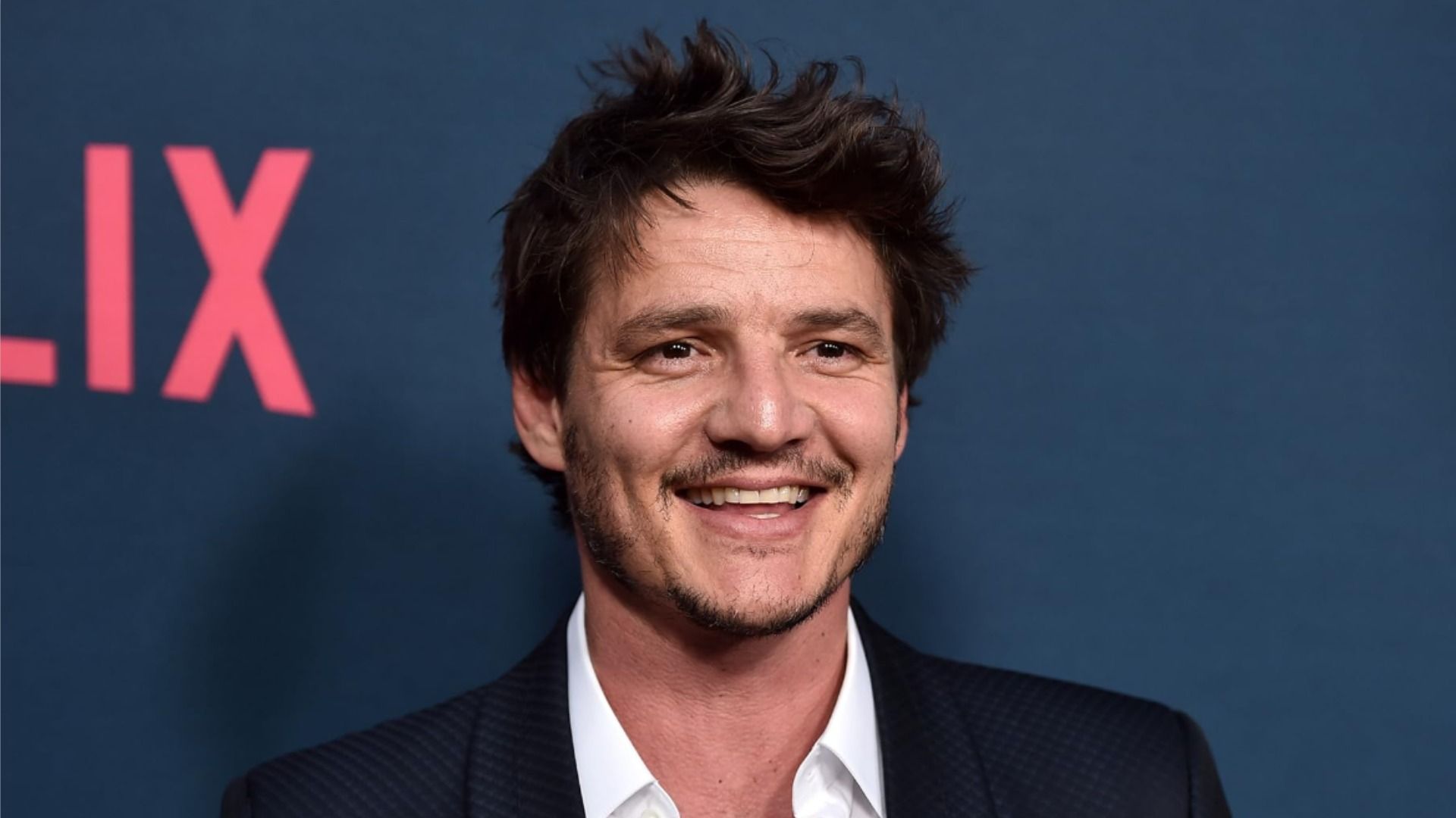 El actor Pedro Pascal con chaqueta negra sonriendo contra un fondo azul
