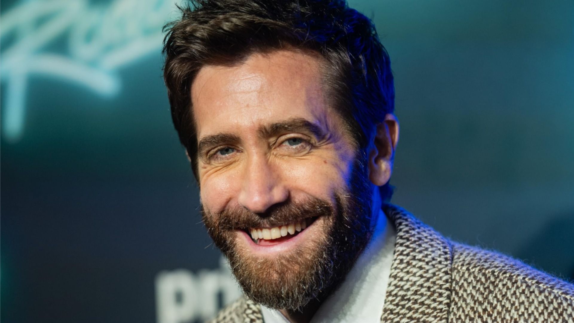 El actor Jake Gyllenhaal con un blazer gris sonríe sobre un fondo azul