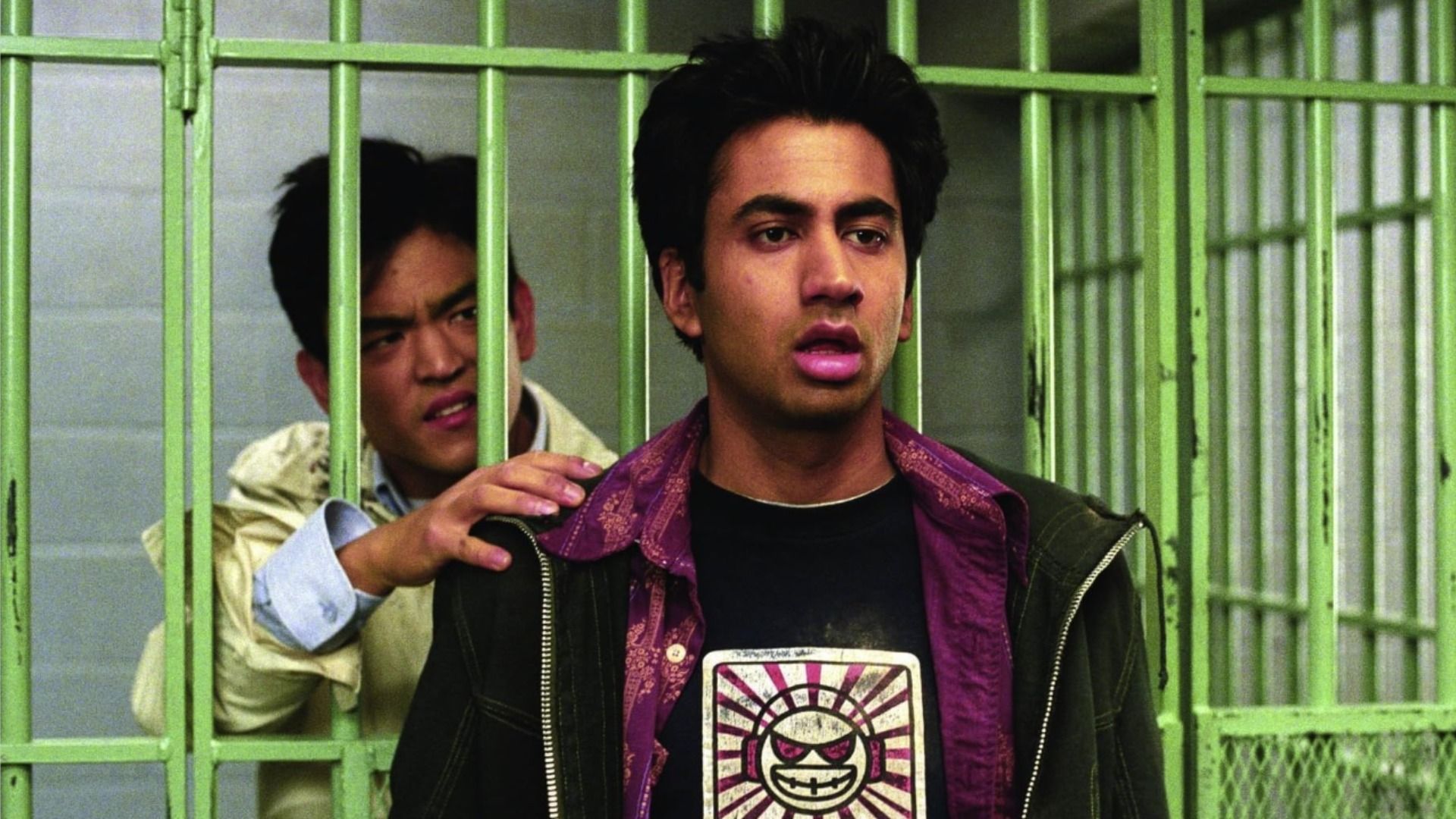 El actor John Cho se para detrás de las rejas verdes de la prisión, el actor Kal Penn se para afuera y mira a un lado