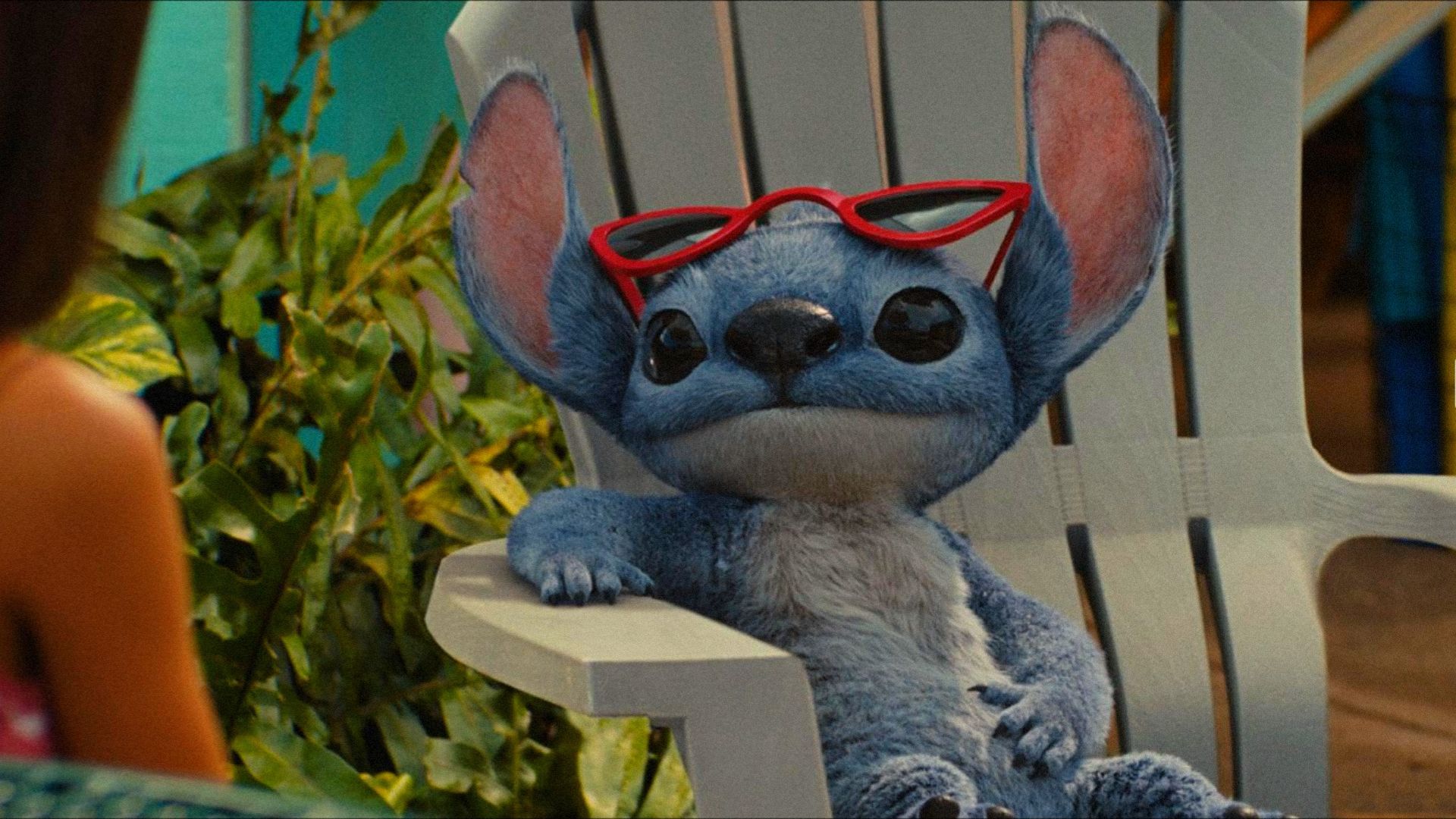 El alienígena azul Stitch se sienta en una silla de playa con gafas y mira hacia un lado