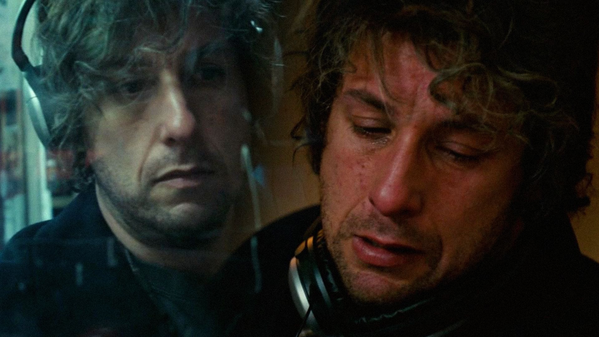 El actor Adam Sandler con pelo rizado está de pie con auriculares y llora