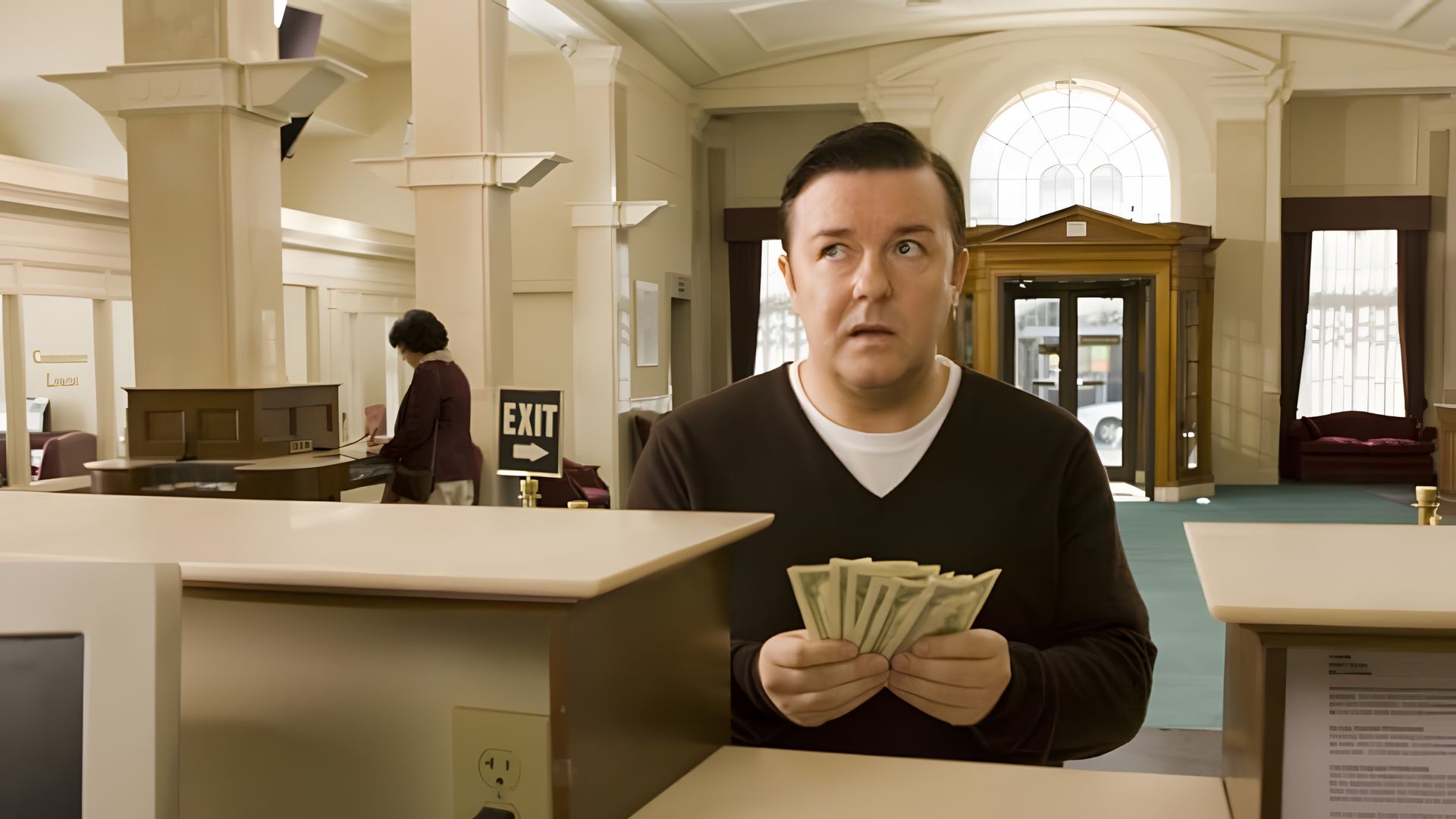Ricky Gervais con expresión de sorpresa sostiene un fajo de dinero en un banco, de pie en el mostrador con una salida y ventanas altas de fondo