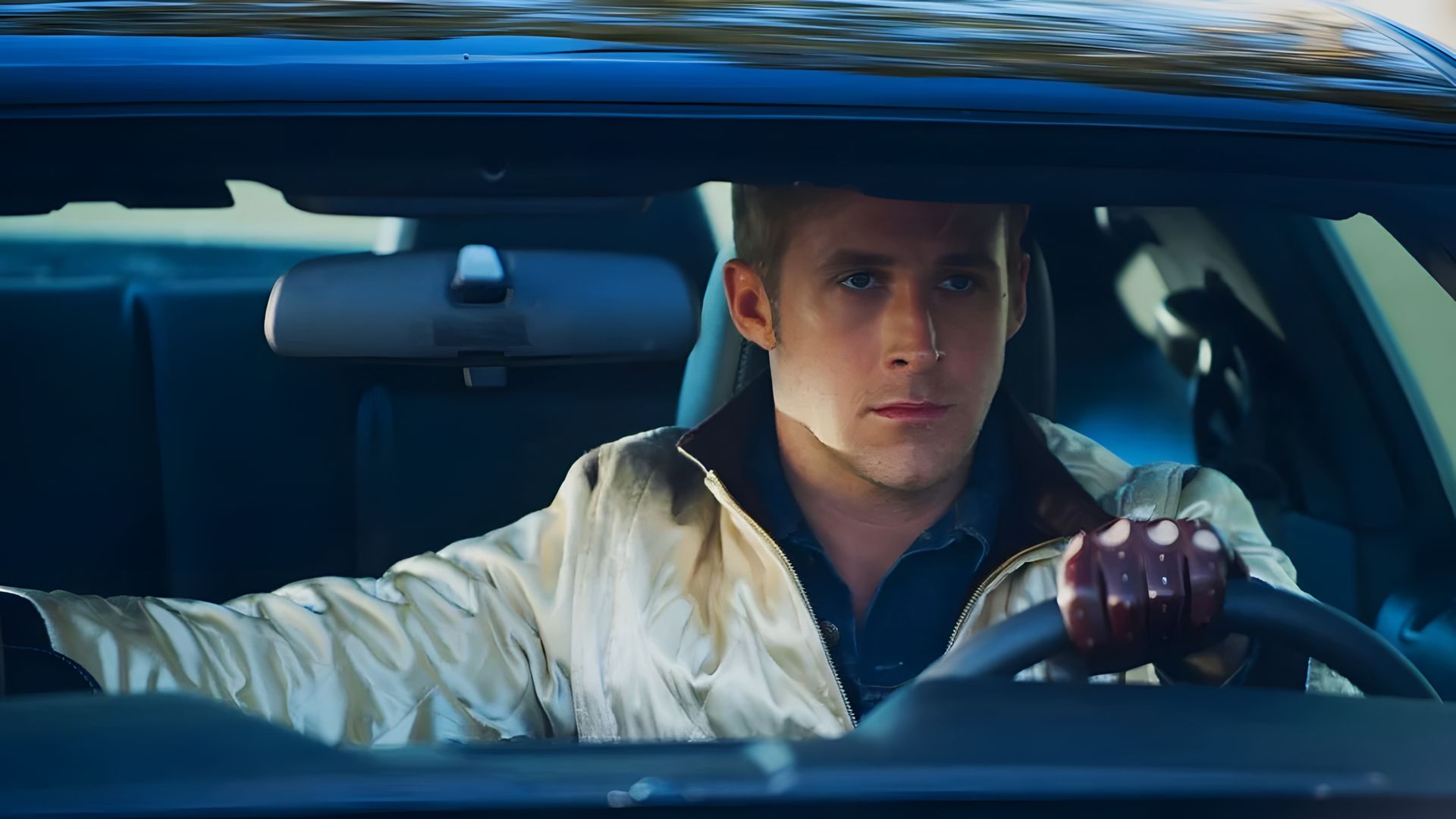 Ryan Gosling con chaqueta de satén y guantes de cuero sentado al volante de un coche, mirando hacia adelante con expresión concentrada