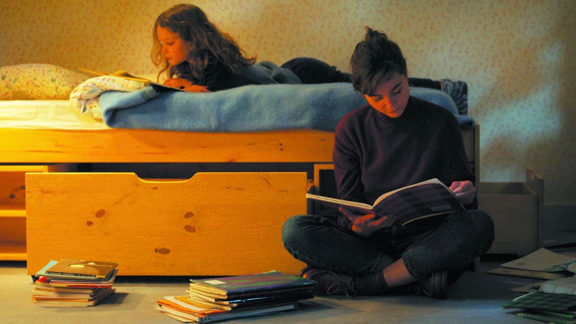 Una niña yace en la cama leyendo un libro mientras una mujer se sienta al lado de la cama con un libro