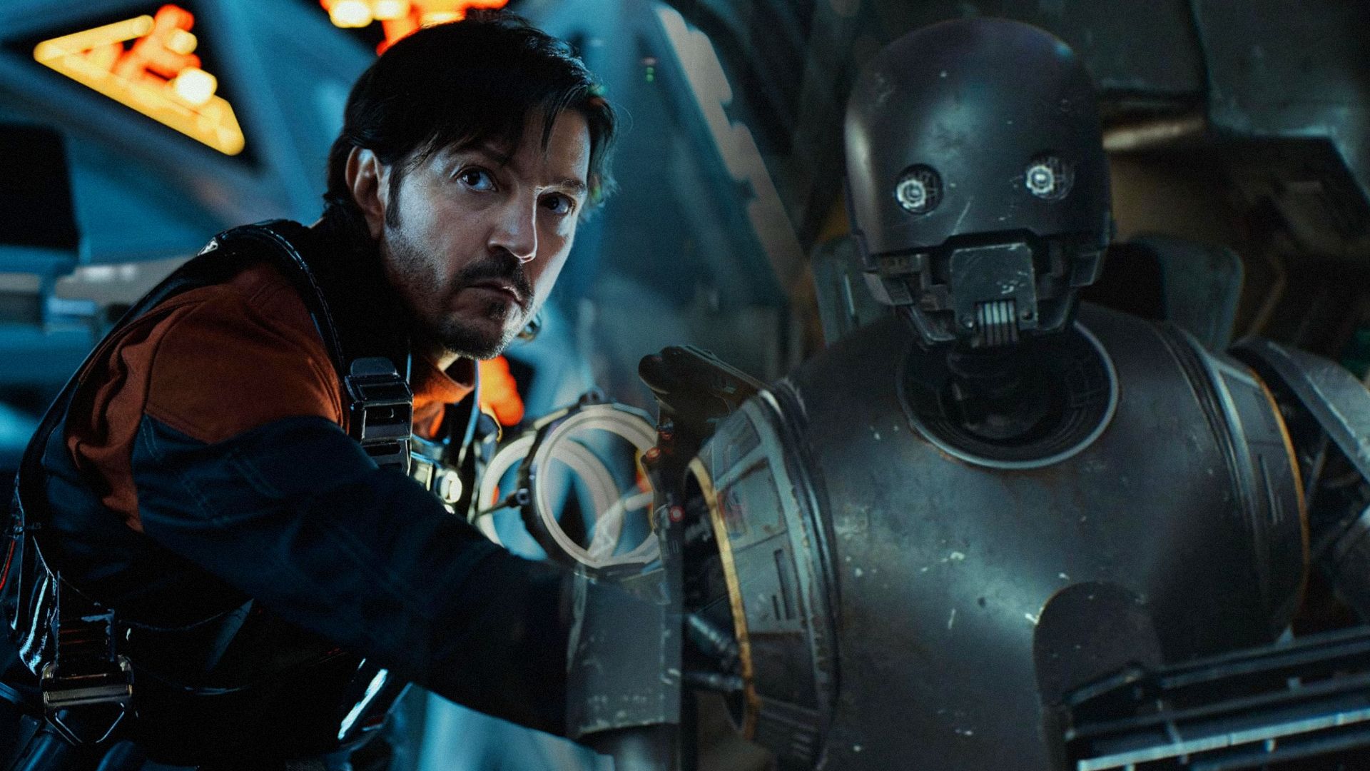 El actor Diego Luna con ropa negra dentro de una nave espacial con un robot gris oscuro cerca