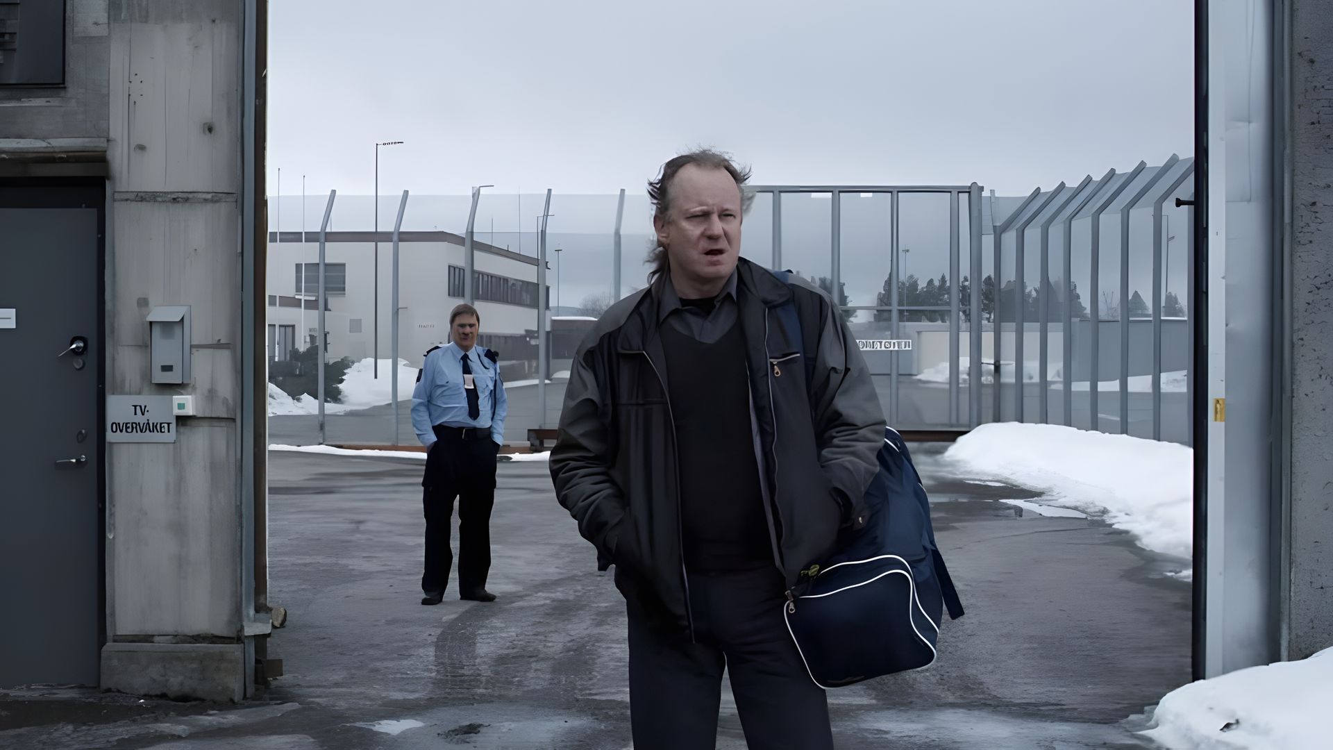Stellan Skarsgård cargando una bolsa sale por las puertas de la prisión, con un guardia de fondo, edificios y nieve visible más allá de la cerca