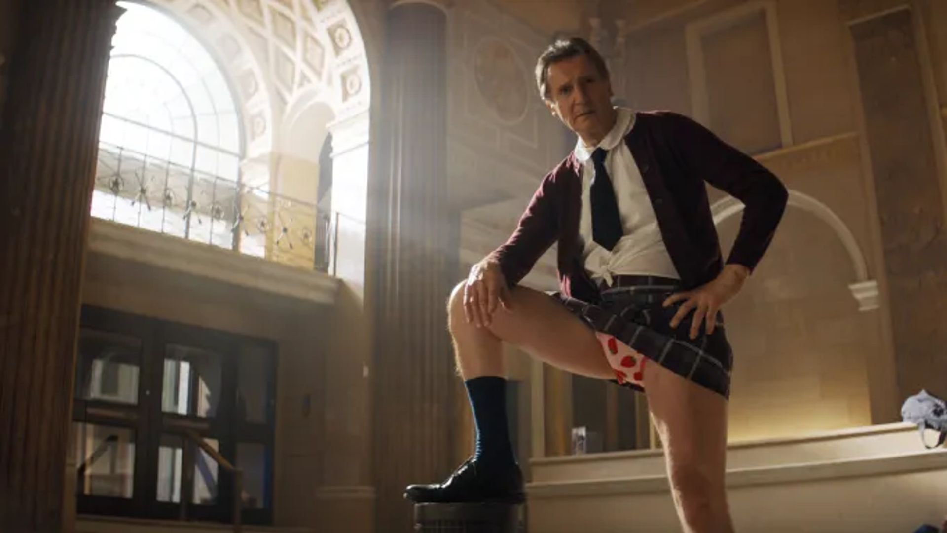 Liam Neeson en uniforme escolar y corbata posa con la pierna levantada, mostrando ropa interior con estampado de corazones, contra un fondo de columnas