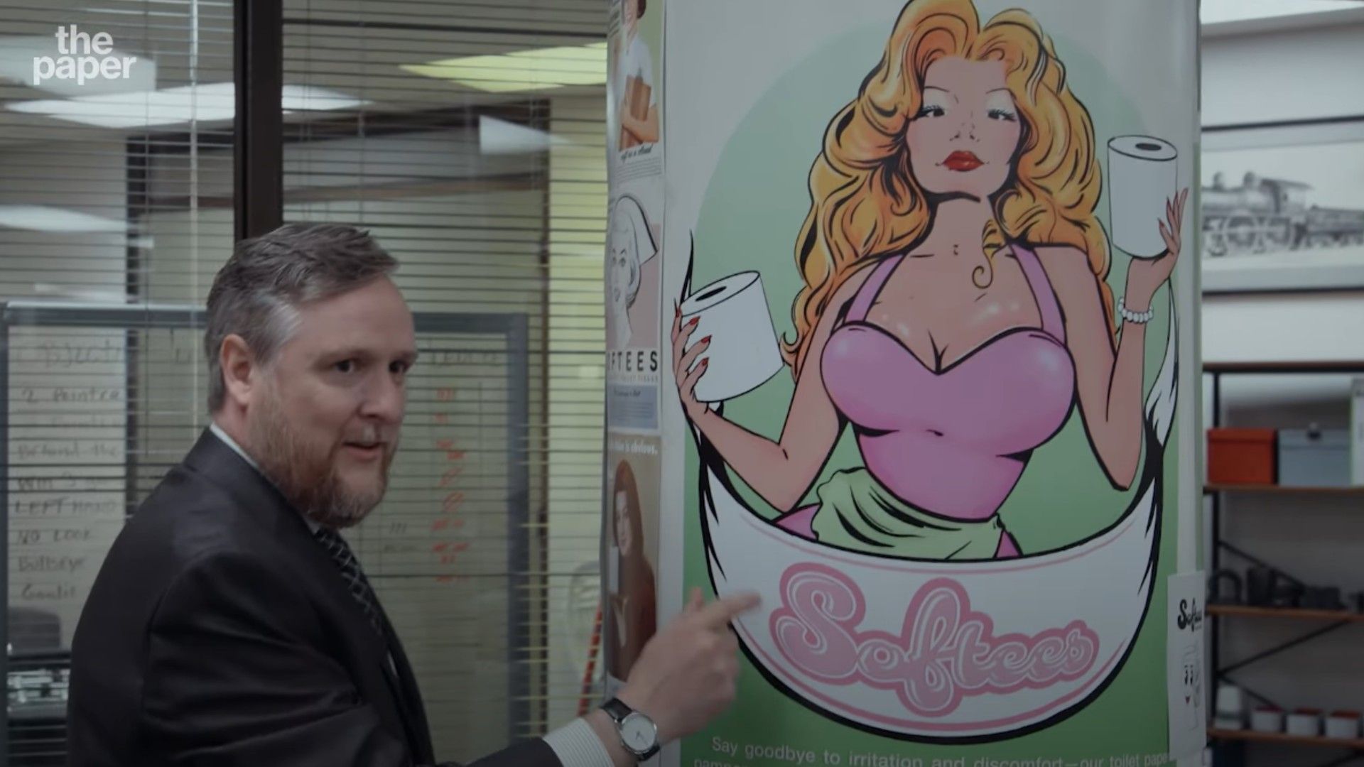 Un hombre en traje junto a un póster con una mujer rubia dibujada sosteniendo rollos de papel higiénico