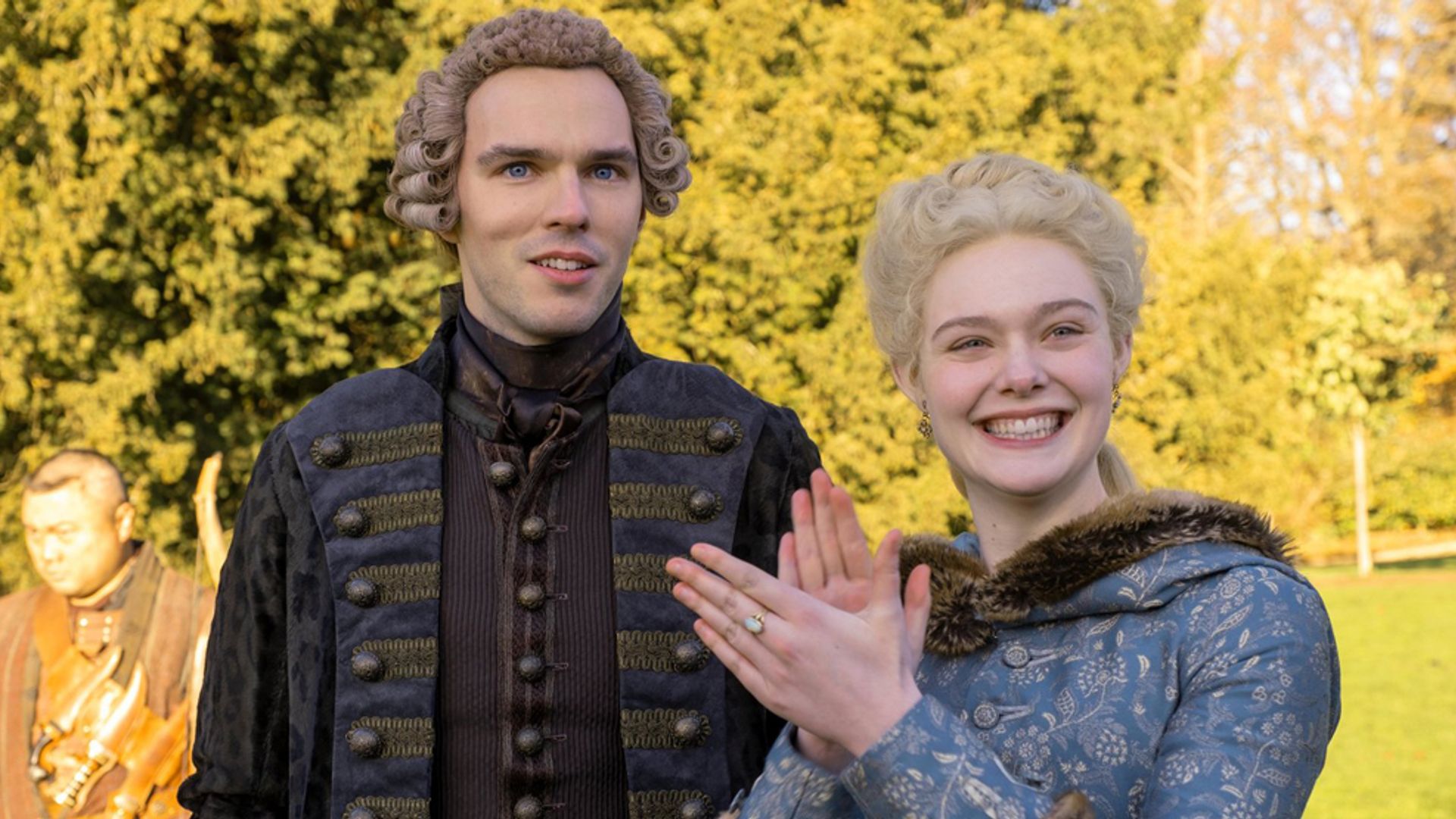 Nicholas Hoult y Elle Fanning con trajes de época del siglo XVIII de pie en la calle, la actriz sonríe y aplaude