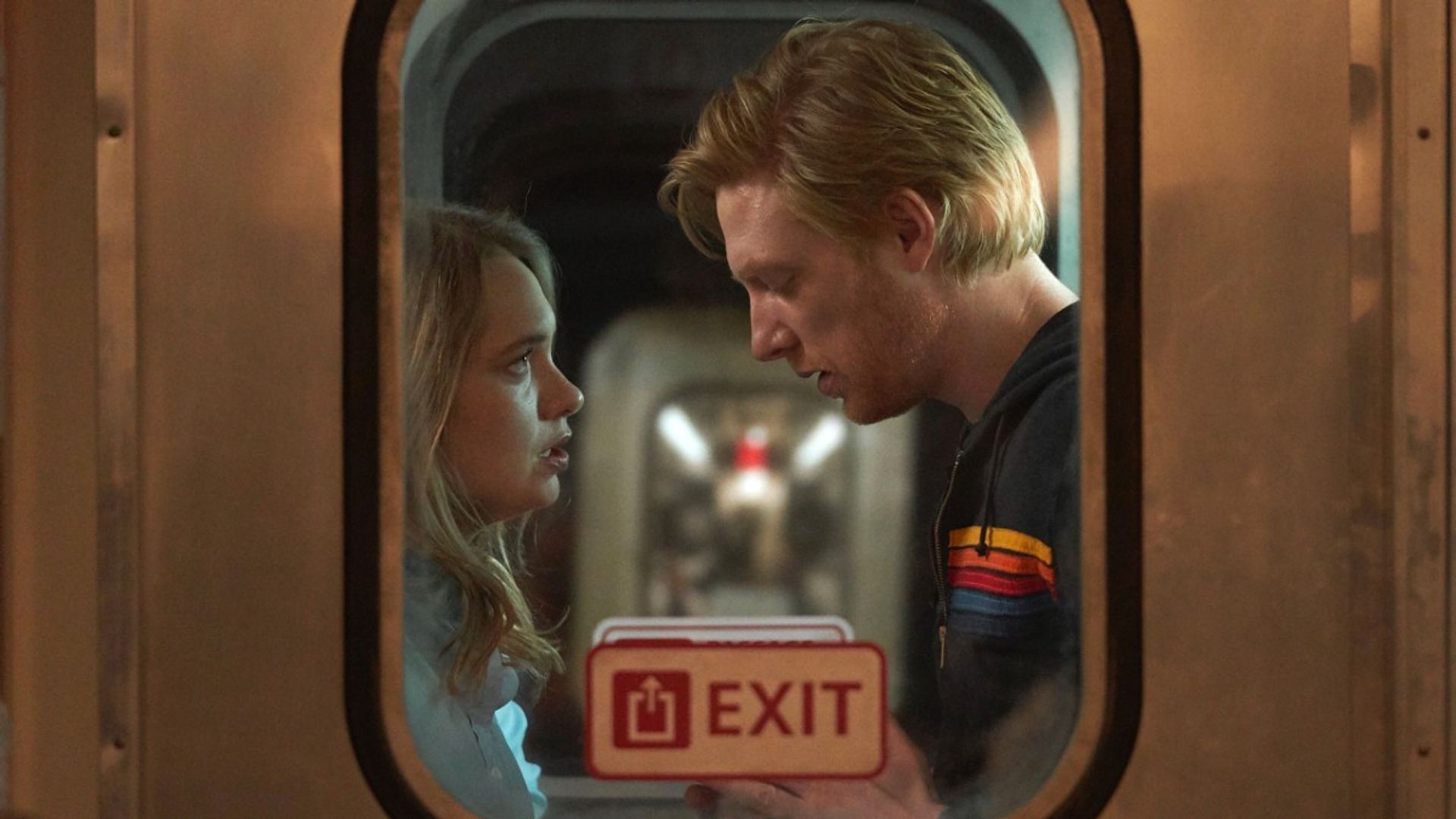 Domhnall Gleeson y Merritt Wever cara a cara en el vestíbulo de un tren, fotografiados desde el exterior de la ventana
