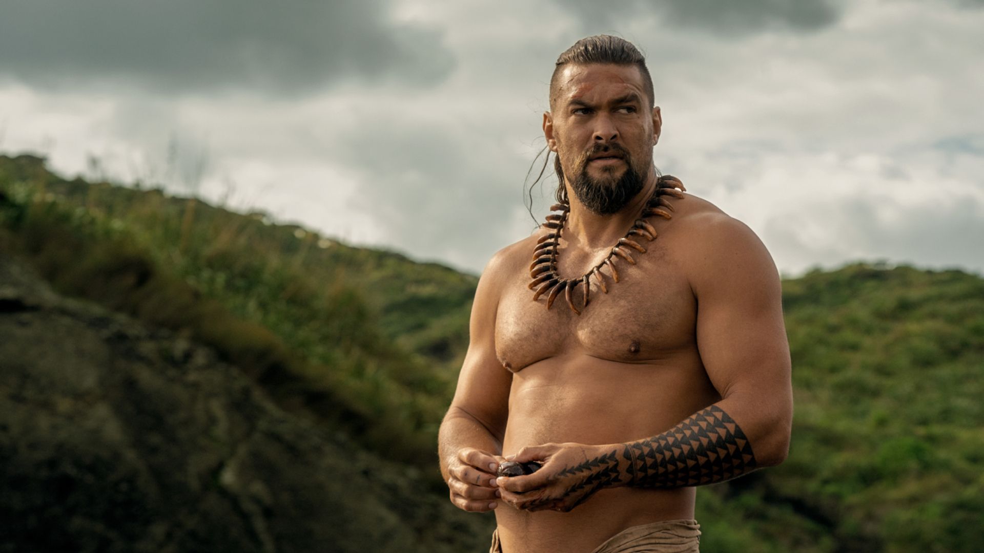 Jason Momoa sin camisa con decoraciones tradicionales hawaianas y tatuajes contra un fondo montañoso
